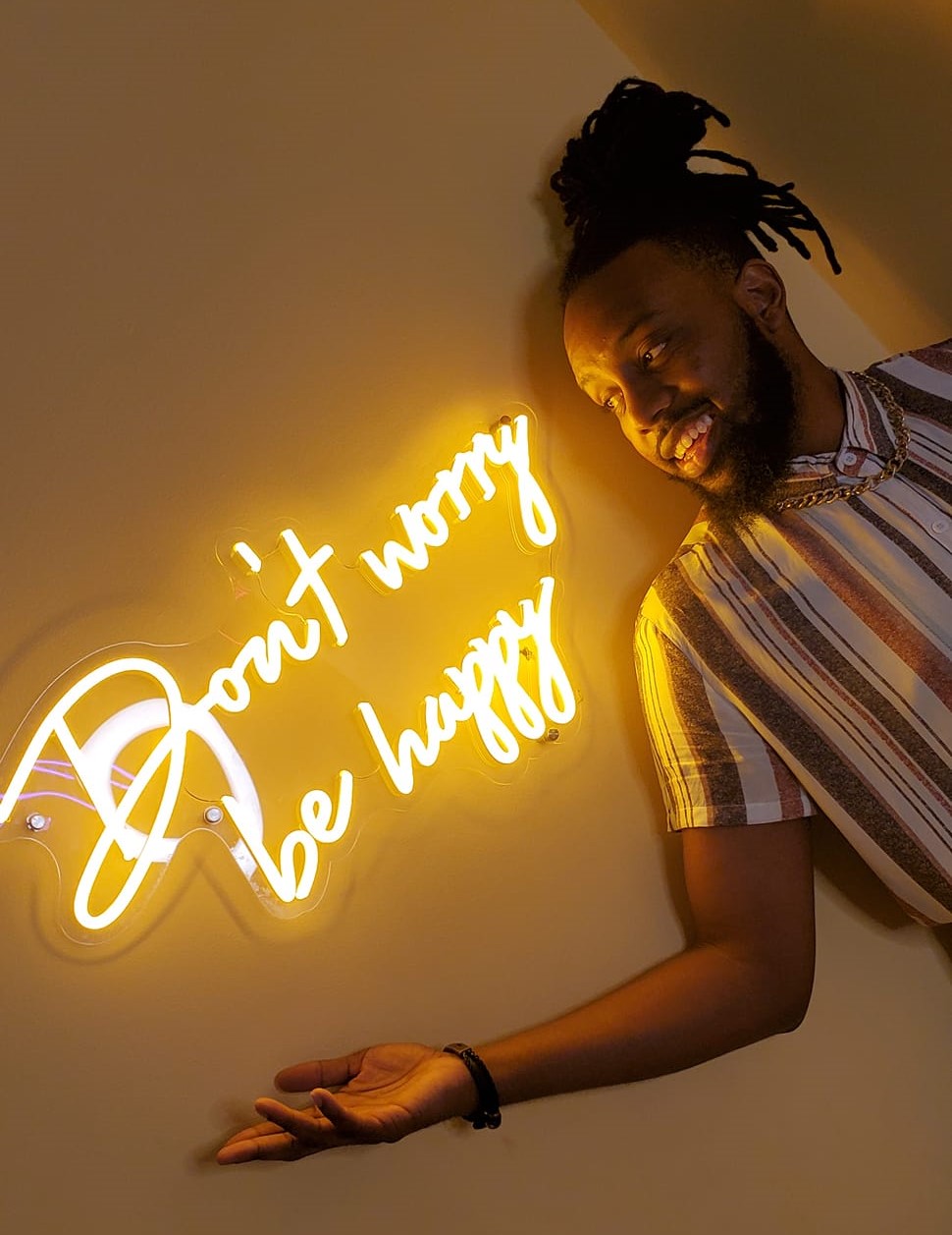 Image of JT Williams smiling next to a neon sign that says "Don't worry be happy"